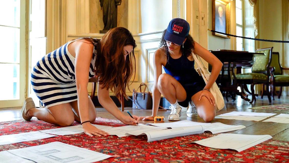 Two women kneel on the floor, intently examining and working on architectural plans spread out on a red carpet in an ornate room.
