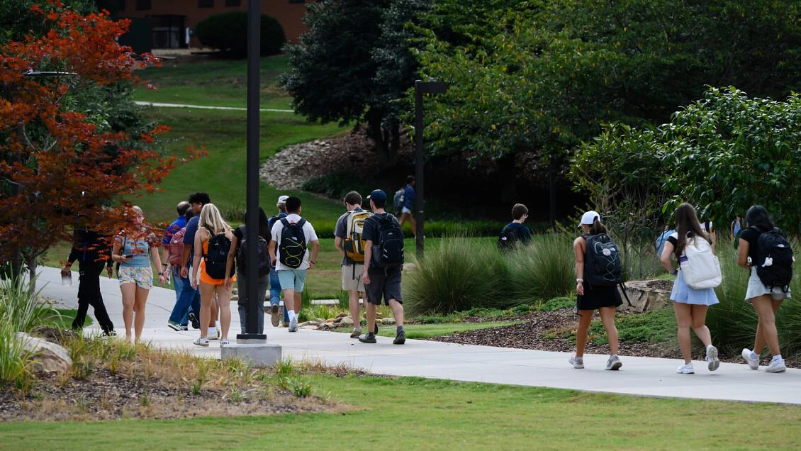 A group of students with backpacks walk along a tree-lined pathway on a sunny day.