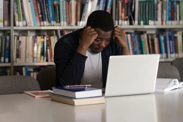 Young man sits at table in library with laptop and a few books, elbows on the table with head resting on hands looking stressed