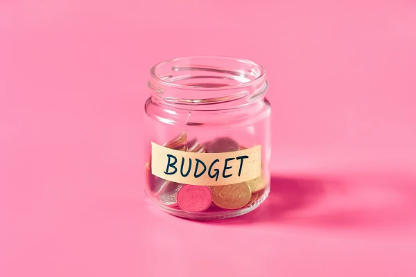 Clear jar about one third full of coins, labeled with tape that reads "Budget", bright pink background