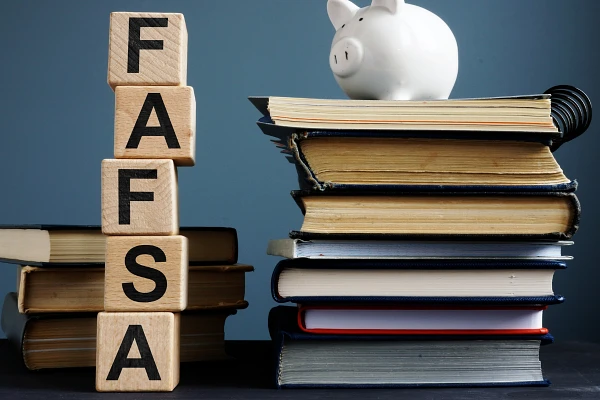 Wooden blocks stacked vertically spelling FAFSA in front of a stack of 3 books and next to a stack of several books and notebooks topped by a white ceramic piggy bank
